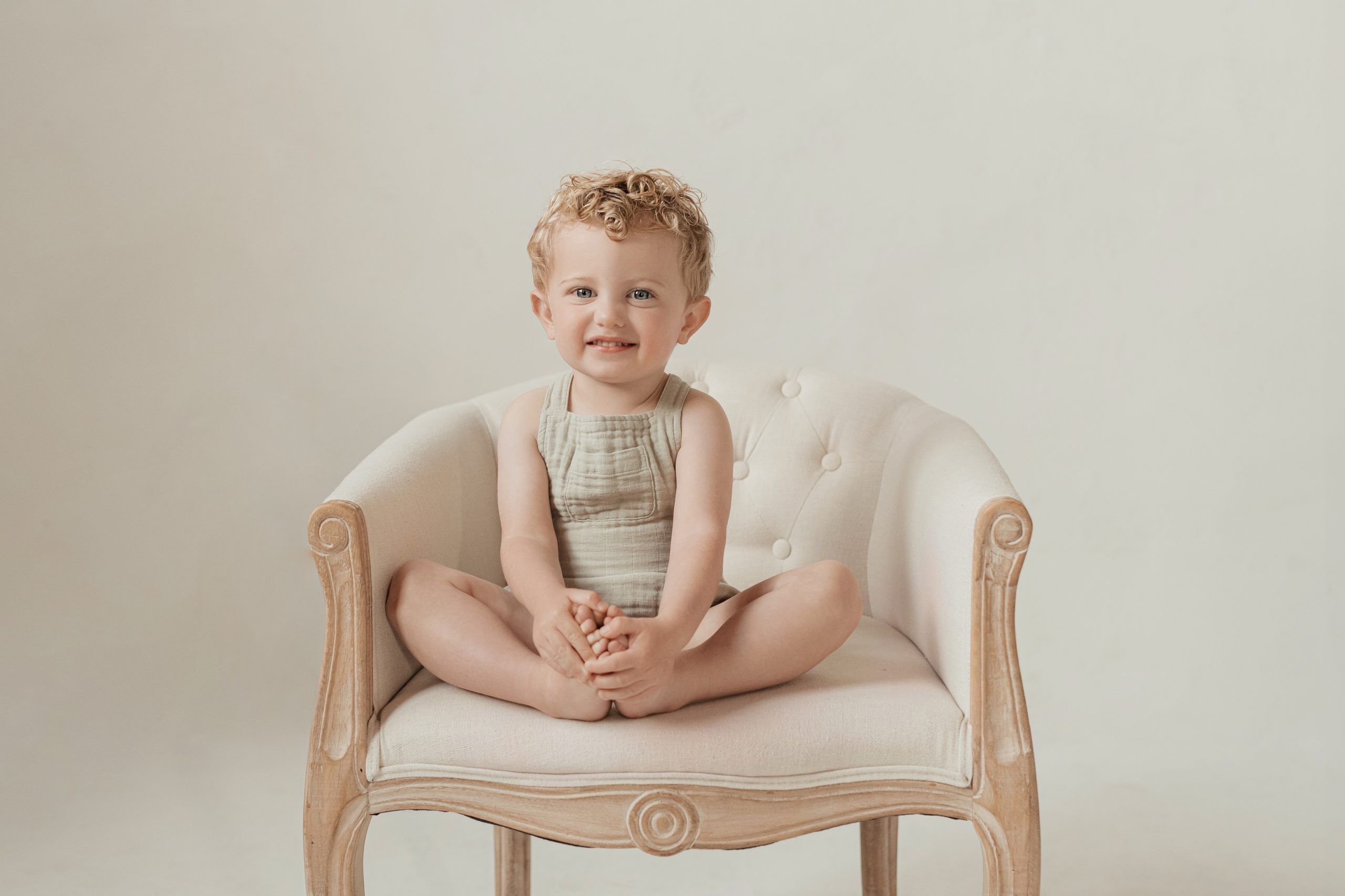little boy with curly hair smiles while sitting on a french style chair in the studio