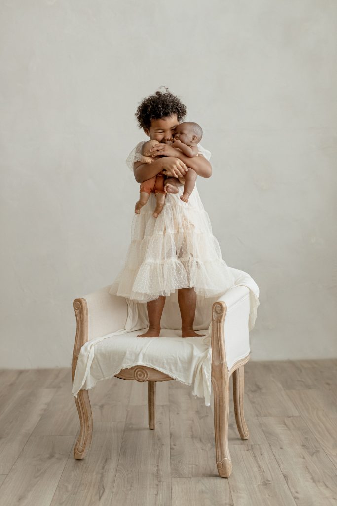 little girl standing on a chair and holding her baby dolls in a photo studio