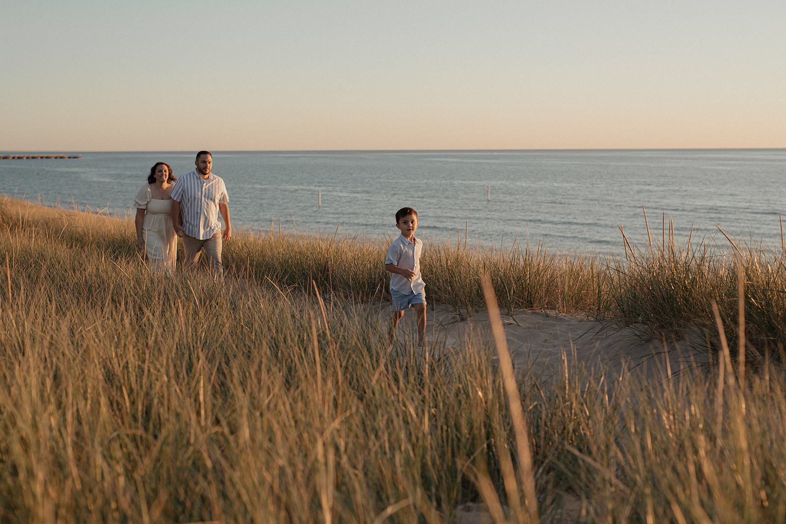 Family photo by Alissa Saylor Photography in the dunes at Windsnest Park, with a young boy running ahead of his parents along a sandy trail overlooking Lake Michigan.