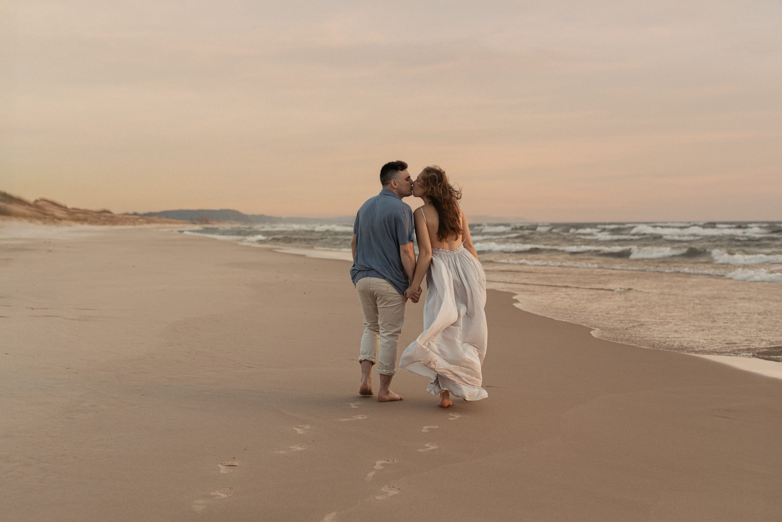 A pregnant mother and father walking along the sandy shoreline of Lake Michigan at Ludington State Park during a peaceful maternity session at sunset.