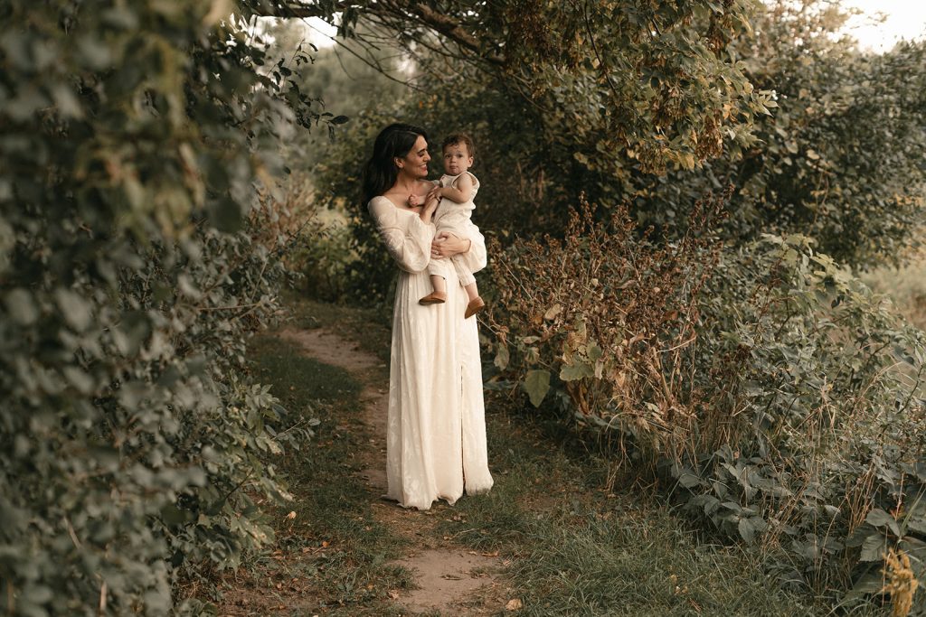 Portrait by Alissa Saylor Photography of a mother holding her baby in the quiet tree line at Roselle Park in Ada, MI, surrounded by greenery and soft light.