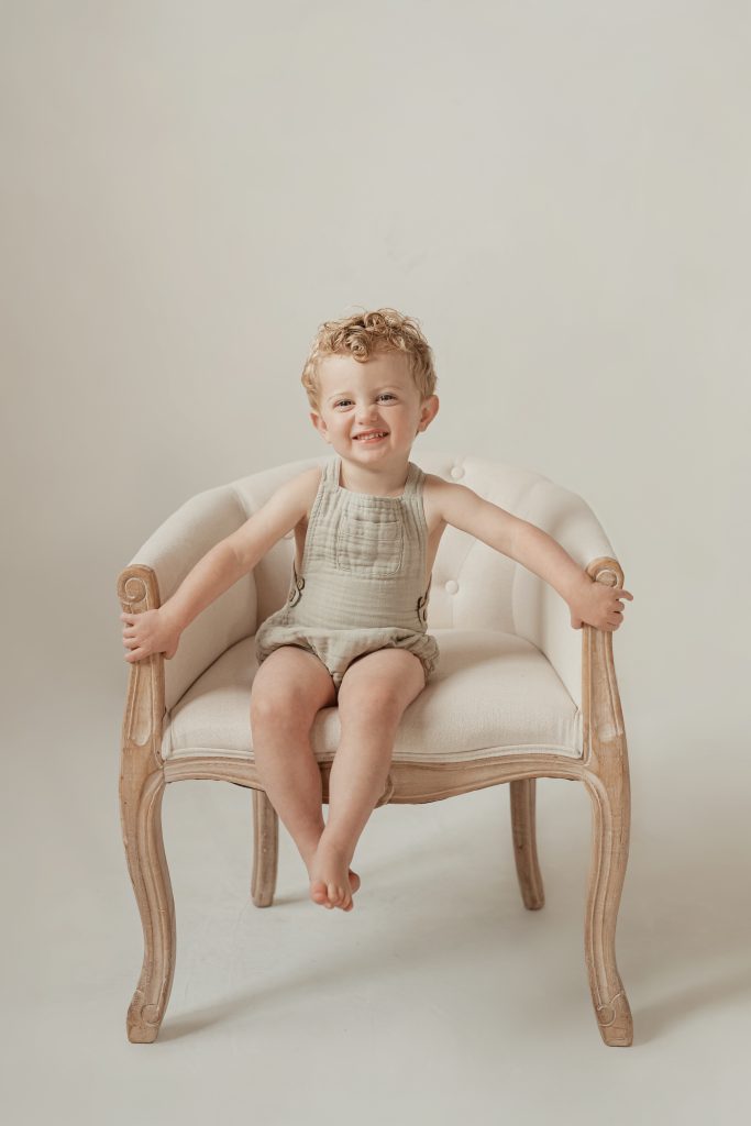 Studio portrait by Alissa Saylor Photography of a smiling 3-year-old boy in a green romper, sitting on a chair in natural light.