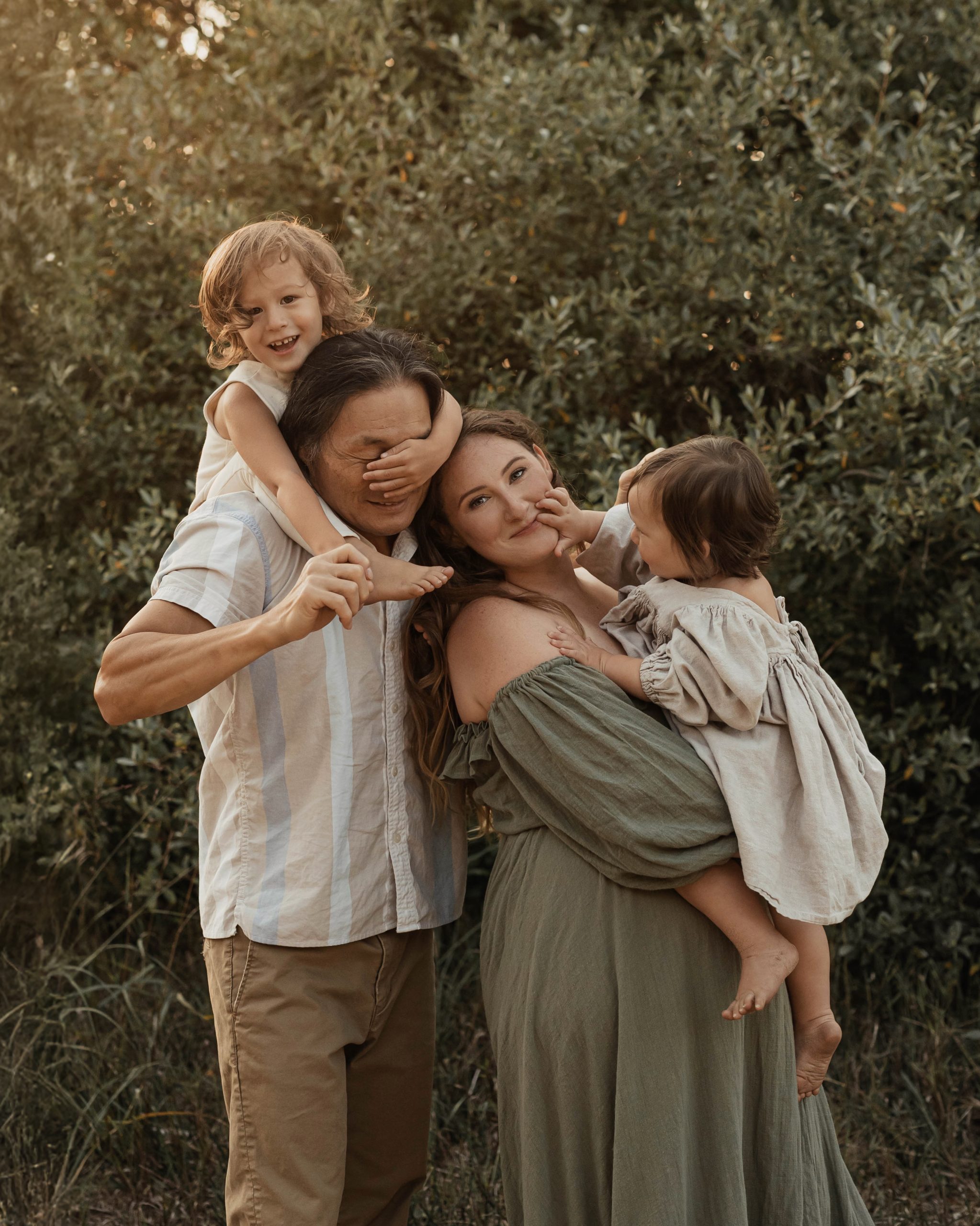 A joyful family of four surrounded by greenery at Lake Michigan. A young child playfully covers dad’s face while sitting on his shoulders, and mom smiles as she holds a baby girl who reaches for her face. Both parents are laughing, capturing a fun and carefree moment.