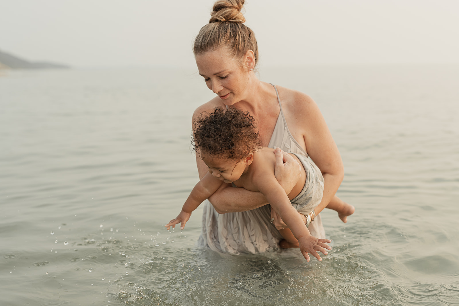 Mother holding her toddler son in the water near Traverse City, captured by Alissa Saylor Photography; she’s wearing a light grey dress as he splashes playfully.