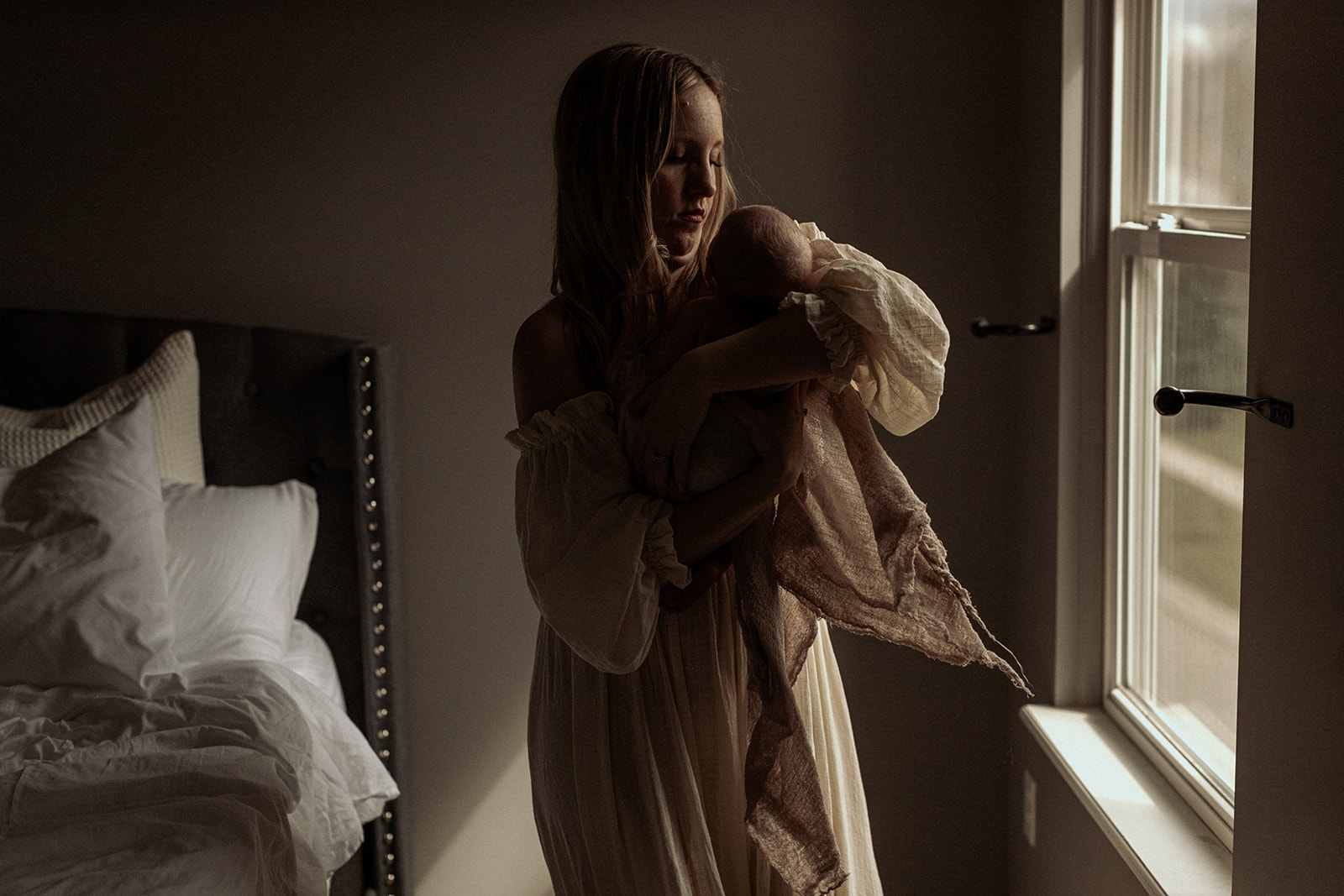 Mother bouncing newborn baby in her bedroom in dark light during an in-home baby session.